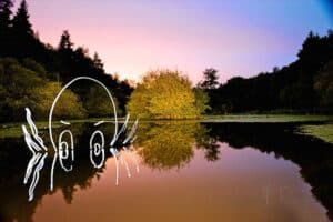 A serene pond surrounded by trees reflecting the sunset sky, poised at twilight. A whimsical white drawing of a ghostly figure's face with wide eyes and hands peeking out from the water, created through light painting photography, adds a playful element to the tranquil scene.