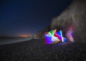 A night-time beach scene with tall, rugged cliffs in the background. The sky is clear and starry. Bright, multicolored light trails form abstract shapes on the pebbled shore, creating a captivating contrast against the dark surroundings—a perfect example of light painting photography.