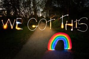A path in a forest is illuminated by light painting photography that reads "WE GOT THIS" above a glowing rainbow. The scene is set during dusk, with trees silhouetted against the remaining light of the sky.