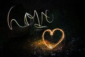 A nighttime scene captured using light painting photography, with the word "hope" and a heart shape both written in the air using sparkling light trails against a dark, leafy background.