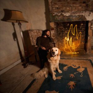 A person sits in an armchair in a dimly lit room with a brick fireplace. A golden retriever stands on a blue rug, with the fireplace artfully illuminated by a yellow neon light shaped like flames, evoking the allure of light painting photography. A tall lamp stands beside the armchair.