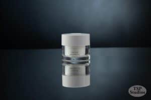 A glass jar of Luminaskin Cream Absolute is centered on a reflective, dark surface against a dark gradient background. The jar is clear with a white lid, and the product name is clearly visible on the label. Expertly crafted cosmetics brand photography by TSP Studios adorns the bottom right corner with their logo.
