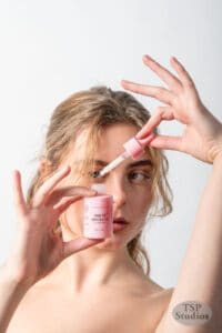 A person with light brown hair holds up a pink bottle labeled "Serum Absolute" and a dropper in front of their face against a plain white background, exemplifying cosmetics brand photography. They appear to be demonstrating or applying the serum, and the bottle features text detailing the product's usage.