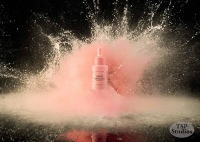 A cosmetic serum bottle with a dropper cap is suspended in mid-air against a dynamic backdrop of pink and white powder explosion. The bottle, captured with stunning cosmetics brand photography, is centered with the words "TSP Studios" at the bottom right corner.