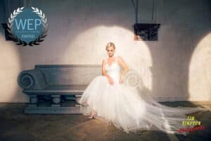 A bride in a flowing white wedding dress sits elegantly on a stone bench. Sunlight highlights her and casts shadows on the wall behind. The image bears the "WEP Award" logo in the top left and "Tim Simpson Photography" in the bottom right corner.