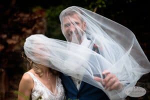 A bride and groom, smiling and partially hidden under the bride's veil, enjoy a candid moment. The groom is in a blue suit, while the bride is in a white lace dress. The background is blurred with greenery, and there is a logo in the corner that reads "Tim Simpson Photography.