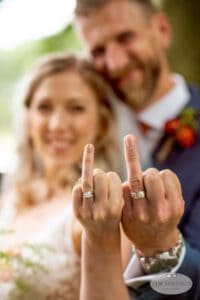 A bride and groom pose for a wedding photo, smiling and holding up their ring fingers to highlight their wedding rings. The background is blurred, and the focus is on their hands and rings. The image is watermarked with "Tim Simpson Photography.
