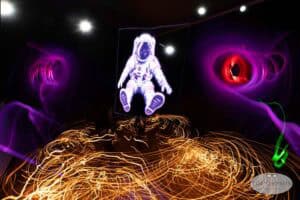 A glowing astronaut illustration floats in a dark room with swirling yellow, purple, and red light trails, evoking the surreal and vibrant energy of light painting photography in a space-themed scene.