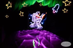 A glowing astronaut stands on a moon-like surface, surrounded by neon stars, a rocket, and swirling green and purple light trails reminiscent of light painting photography against a dark background.