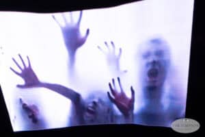Blurred figures with outstretched hands and open mouths press against a frosted glass, creating a dramatic and haunting scene reminiscent of light painting photography.