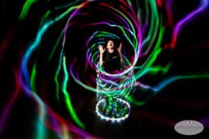 A person stands surrounded by swirling, colorful light trails in a dark space, looking up with hands raised in excitement. The tunnel-like spiral of lights captures the magic of light painting photography.