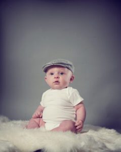 A baby wearing a white onesie and a grey cap sits on a white, furry surface against a plain grey background. The baby looks slightly upward with a curious expression, capturing the essence of family photography.