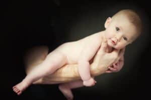 A baby with soft, fair skin is held securely by an adult's hands against a dark background. The unclothed infant looks towards the camera with wide eyes and slightly parted lips, conveying a sense of curiosity and calm—a beautiful moment in family photography.