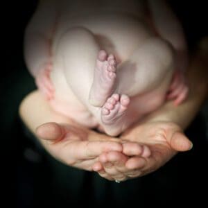 A close-up image of an adult's hands gently cradling a newborn baby's feet. The baby's tiny feet are the focus, with toes slightly curled, while the rest of the baby's body is blurred in the background. The scene emanates warmth and tenderness, capturing a timeless moment in family photography.