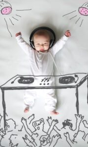 A baby dressed in a white onesie lays on a white surface with arms raised, wearing large black headphones. Drawn-in illustrations show DJ equipment in front of the baby, lights above, and a cheering crowd below, making it look like the baby is DJing—an adorable moment captured in family photography.