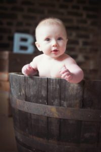 A baby with light hair is standing inside a wooden barrel, looking directly at the camera. The baby is naked and holding onto the edge of the barrel, with a softly focused background that includes a brick wall and a large letter "B". This charming family photography captures the innocence and joy of childhood perfectly.