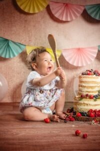 A joyful baby sits on a wooden floor, holding a messy wooden spoon in front of a half-eaten cake adorned with berries. Wearing a floral dress, the baby is surrounded by colorful paper fans and balloons that create a festive atmosphere, perfect for capturing heartwarming moments in family photography.