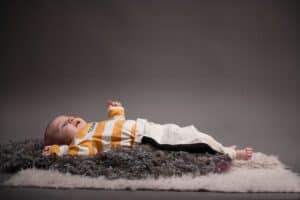 A baby lies on a soft, textured rug against a neutral background. The baby is wearing a yellow and white striped long-sleeve shirt and light-colored pants, looking upward with arms outstretched and feet comfortably resting on the rug.
