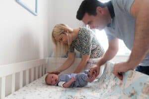 A man and woman smile warmly as they bend over a baby lying in a crib. The baby, dressed in a blue outfit, rests on a patterned crib sheet. The woman gently touches the baby's head while the man adjusts a colorful blanket. This tender moment captures the essence of family photography.