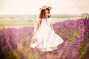 A young girl wearing a white dress and a floral wreath on her head walks through a field of blooming lavender, perfectly capturing the essence of family photography. The scene is bright and colorful, with soft sunlight and a glimpse of a rainbow in the background.