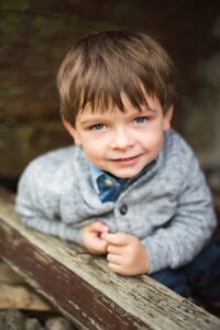 A young boy with short brown hair and blue eyes smiles at the camera. He is wearing a grey knitted cardigan and a blue shirt. His hands are clasped together in front of him, leaning against a worn wooden surface with a blurred background—a perfect moment captured in family photography.