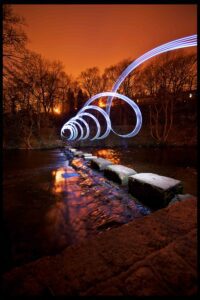A scenic view of a river at dusk, with stepping stones leading across it. Spiraling light trails hover above the river, capturing the essence of light painting photography and creating a dynamic and futuristic effect against the backdrop of silhouetted trees and an orange sky. The reflection shimmers on the water.