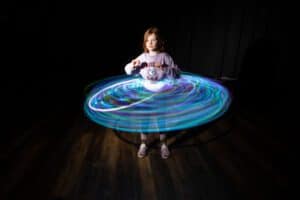 A young girl stands in a dark room, illuminated by colorful LED lights spinning around her, creating a circular light trail effect. Engaging in light painting photography, she holds a piece of the equipment creating the lights, which appears to be a hula hoop. She is wearing a light-colored sweatshirt and shoes.