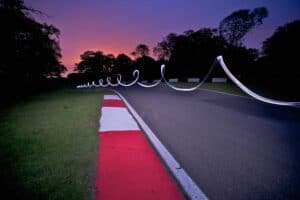 A long-exposure photo of a racetrack at twilight showcases light painting photography, capturing a curving trail of white light hovering above the track. The red and white curb on the left contrasts with the gradient sky from purple to orange, while silhouetted trees line the background.