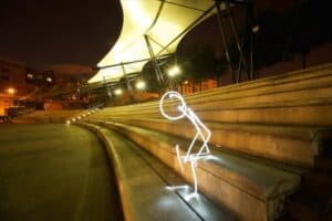 A nighttime scene features an illuminated, glowing stick figure, created through light painting photography, sitting on stone steps under a modern, tent-like canopy structure. The area is softly lit, with surrounding buildings and trees dimly visible in the background.