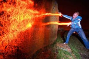 A person dressed in outdoor clothing appears to shoot bright orange light beams from their hands towards a large rock at night. The scene, reminiscent of light painting photography, is dramatic with an illuminated rock face and dark surroundings, giving it an energetic and fantastical feel.
