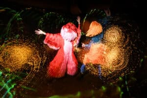 Two people in colorful, fringed costumes dance energetically under swirling, golden light trails, creating a vibrant and dynamic scene with motion and festive atmosphere—perfectly capturing the essence of light painting photography.