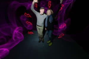 An older man and woman stand smiling and waving with one arm raised. The background is dark, illuminated by swirling purple light and red lightning bolt shapes—captured in the dramatic style of light painting photography.