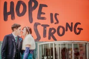 A couple, dressed in formal attire, stands closely together, smiling in front of a bright red background with large black text that reads, "HOPE IS STRONG." The woman is wearing a white fur stole and long gloves, while the man is in a dark suit with a blue boutonniere.