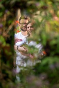 A couple stands in a garden, smiling and holding a newborn baby, framed by leafy green foliage in the foreground. The man wears glasses and a white shirt, while the woman also wears a white shirt. Red flowers can be seen in the background, capturing the essence of family photography.