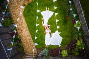 An overhead view of two brides in white wedding dresses lying on a green lawn in a garden. They are holding hands while surrounded by white triangular pennant banners and garden pathways. The scene is colorful and celebratory.