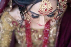 Close-up shot of a bride in traditional South Asian attire. Her intricate jewelry and headpiece, adorned with red and gold details, compliment her embellished gold dress. The image is taken from above, highlighting her makeup and long eyelashes as she looks down.