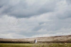 A couple in wedding attire stands closely together on a grassy field near a stone wall under a cloudy sky. A flock of birds is flying in the distance. The landscape is vast and open, giving the image a serene and expansive feel.
