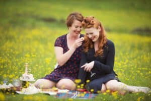 Two women sit on a picnic blanket in a field of yellow flowers in Oldham, enjoying a moment perfect for family photography. One woman playfully places a flower in the other's hair, surrounded by picnic items like champagne, glasses, and various foods. Both are smiling and savoring the moment.