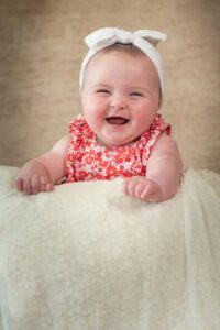 A smiling baby wearing a white headband with a bow and a red floral dress is lying on a soft, cream-colored blanket. The baby has rosy cheeks and is looking directly at the camera, radiating joy. The background is a neutral beige color.