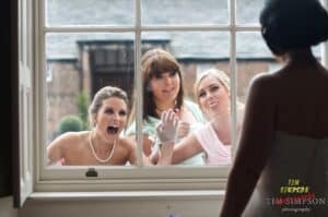 Three women outside a window making funny faces and gestures towards a person facing them inside. The window creates a barrier showcasing playful interaction. Building exterior is visible in the background.
