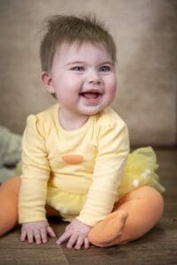 A happy baby with light hair sits on a wooden floor. The baby is dressed in a yellow onesie with a skirt and orange polka-dotted tights. The baby is smiling with an open mouth and looking slightly to the side.