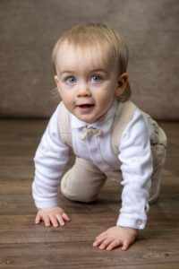 A young child with light hair and blue eyes is crawling on a wooden floor. The child is wearing a white long-sleeved shirt, beige suspenders, and pants. The background is out of focus with a neutral tone.