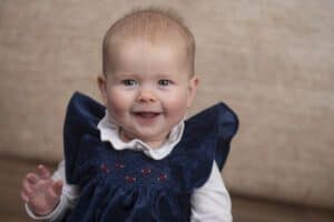 A smiling baby with light hair sits against a beige background. The baby is wearing a blue velvet dress with intricate red stitching and a white-collared shirt underneath, while reaching out with one hand.