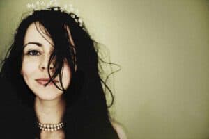 Close-up of a woman with long, dark hair partly covering her face, wearing a delicate floral headpiece and a beaded necklace. She is smiling slightly against a plain, muted background.