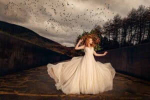 A woman with red hair and a flower crown stands in the middle of a desolate path with barren trees on either side. Wearing a flowing, cream-colored gown, she strikes a dynamic pose. Birds fill the cloudy sky above her, adding to the dramatic scene.