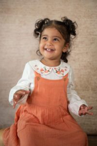 A young child with curly hair, styled in pigtails, smiles brightly. They are wearing a white long-sleeved shirt with floral embroidery on the collar and an orange pinafore dress. The background is a neutral beige.