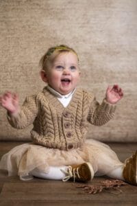 A smiling baby wearing a beige knitted sweater, white tights, brown shoes, and a light-colored tutu sits on a wooden floor in front of a beige background. The baby has a thin headband adorned with small flowers and has both arms raised joyfully.