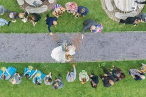 Aerial view of a wedding ceremony. The bride in a white dress and the groom in a light gray suit walk down a gravel path, while guests on both sides throw confetti. The green lawn is dotted with wooden circular tables.