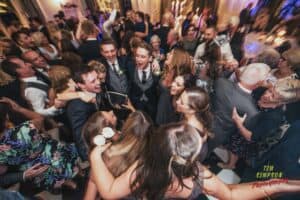 A lively group of people huddle together in a circle at a festive event, smiling and singing. The crowd is dressed in formal attire, with men in suits and women in dresses. The room is decorated with elegant lighting and filled with other guests enjoying the celebration.