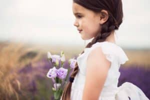 A young girl with long, dark braids and a white dress stands sideways in a field of lavender flowers, holding a small bouquet of light purple blooms. The background is slightly blurred, showcasing the field and giving the image a dreamy, peaceful feel—perfect for family photography.
